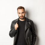 Handsome bearded man expressing confidence, touching his leather jacket and looking self-assured, white background.